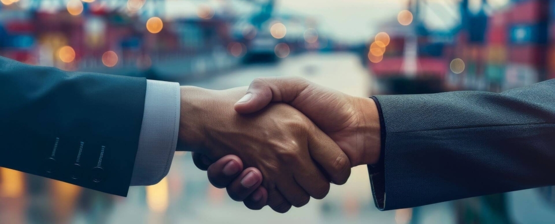 2 people shaking hands, edges of their suit arms visible against maritime backdrop